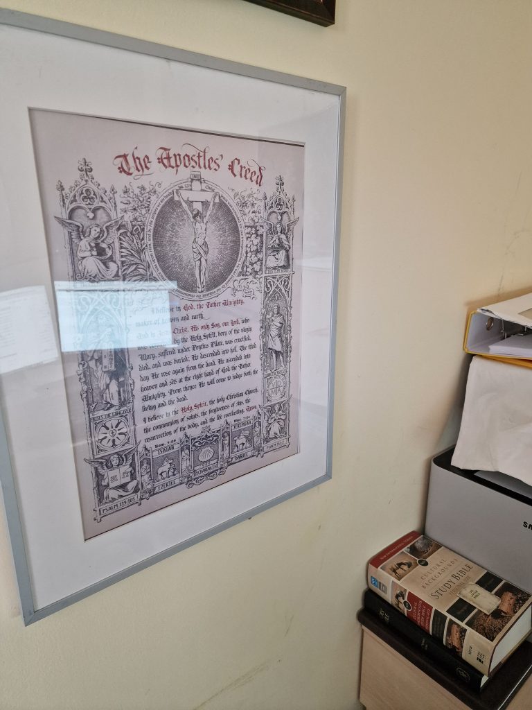 Apostles’ Creed Framed In Office – Doctrine Matters Framed print of the apostles’ creed hanging on a home office wall above a stack of bibles, symbolising the personal importance of historic christian doctrine.